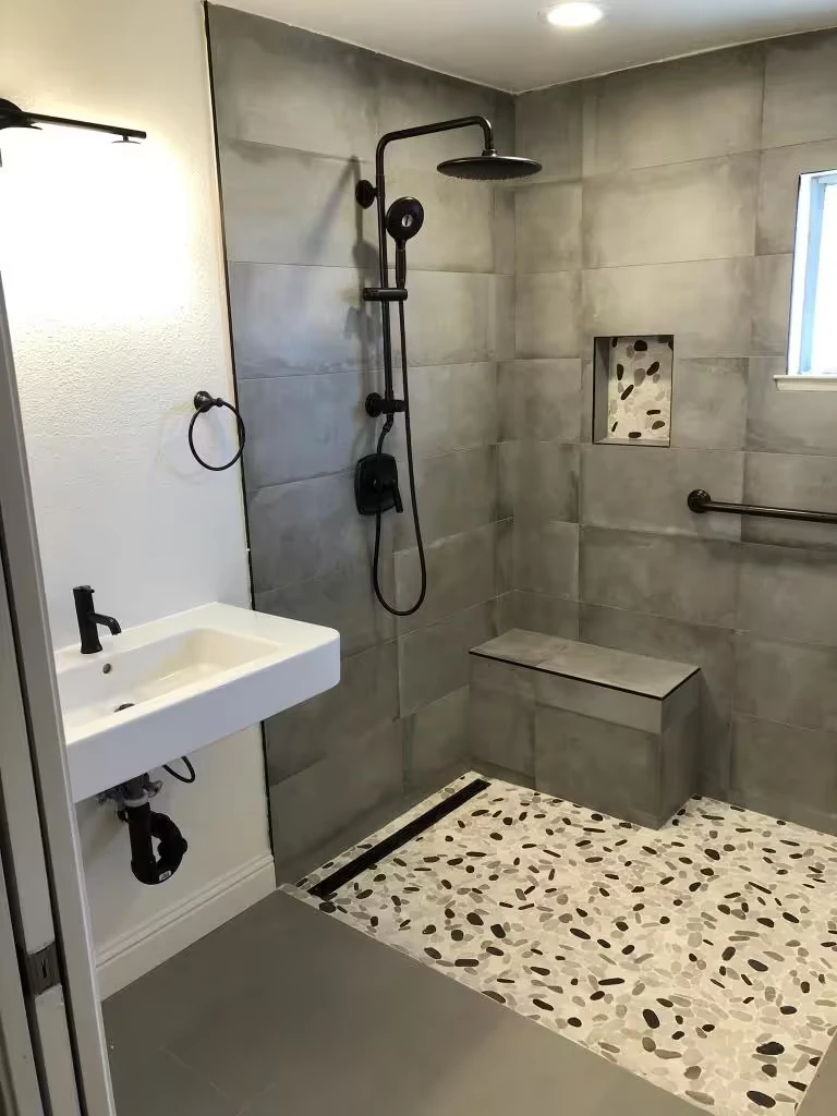 Modern bathroom with marble walls and floor, glass shower enclosure, wooden vanity with white sink, toilet, and decorative plants beside a wooden door.