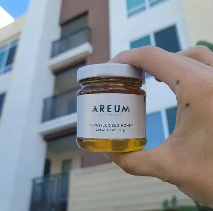 A hand holding an Areum Apartments branded unpasteurized honey jar held up in front of an apartment building on a sunny day