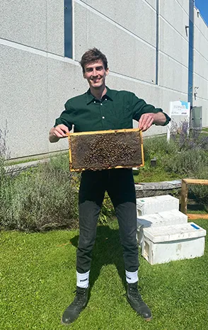 Man holding a honeycomb frame teeming with bees while standing in front of a suburban building and beehive, Alvéole return-to-office program