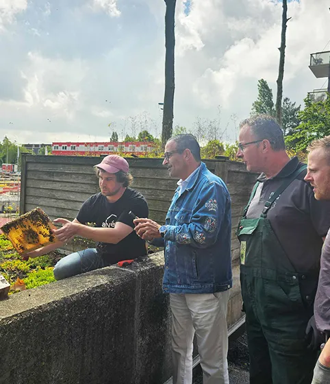 Alvéole beekeeper presenting a honeycomb frame to three attentive visitors in a rooftop garden, return-to-office program