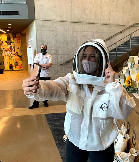 Woman in Alvéole beekeeping gear trying on a veil and holding a smoker indoors at a JLL building activation