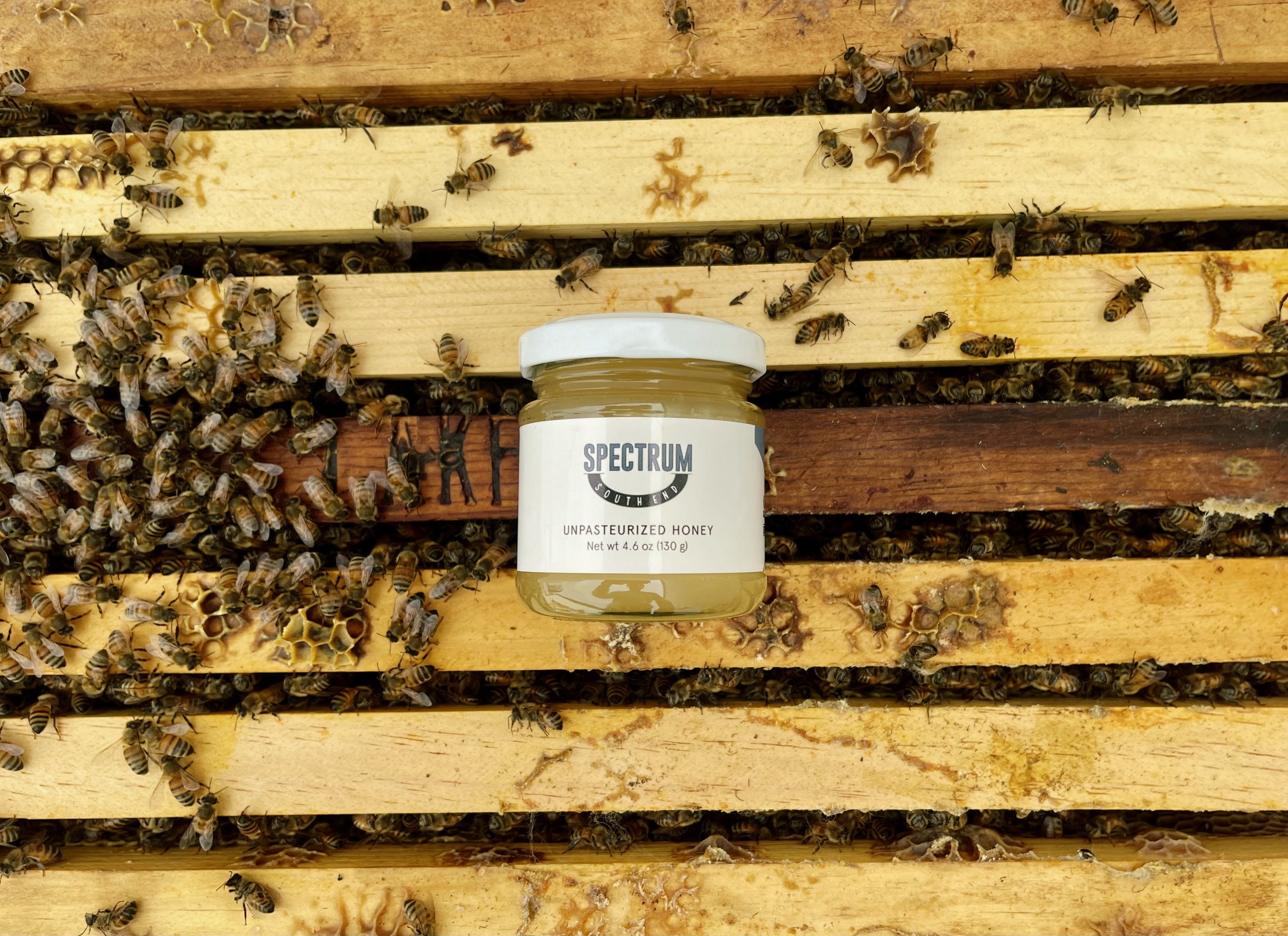 A jar of Spectrum unpasteurized honey resting on a beehive frame surrounded by live bees