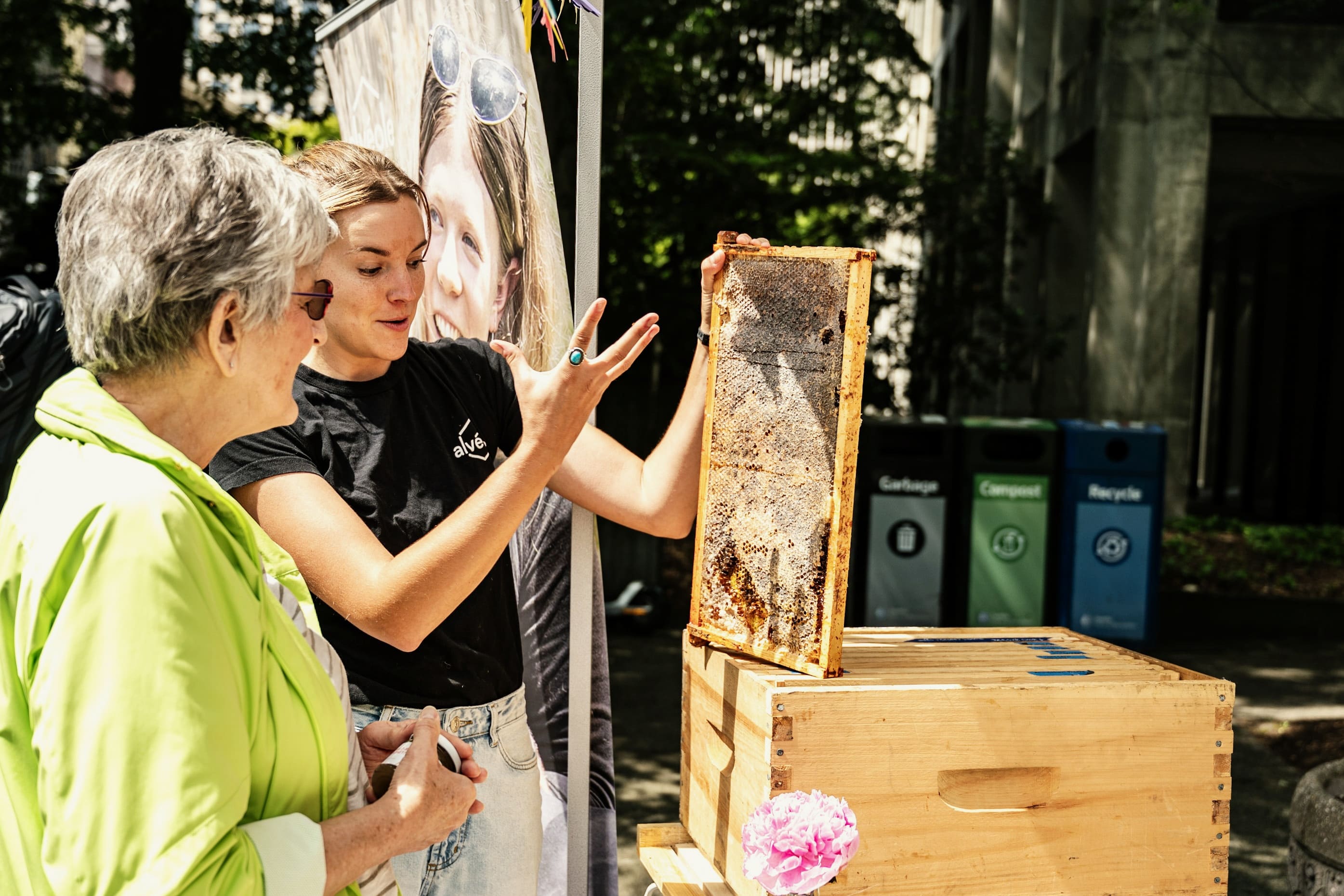 An Alveole beekeeper showing a honeycomb frame to a woman at an outdoor event table