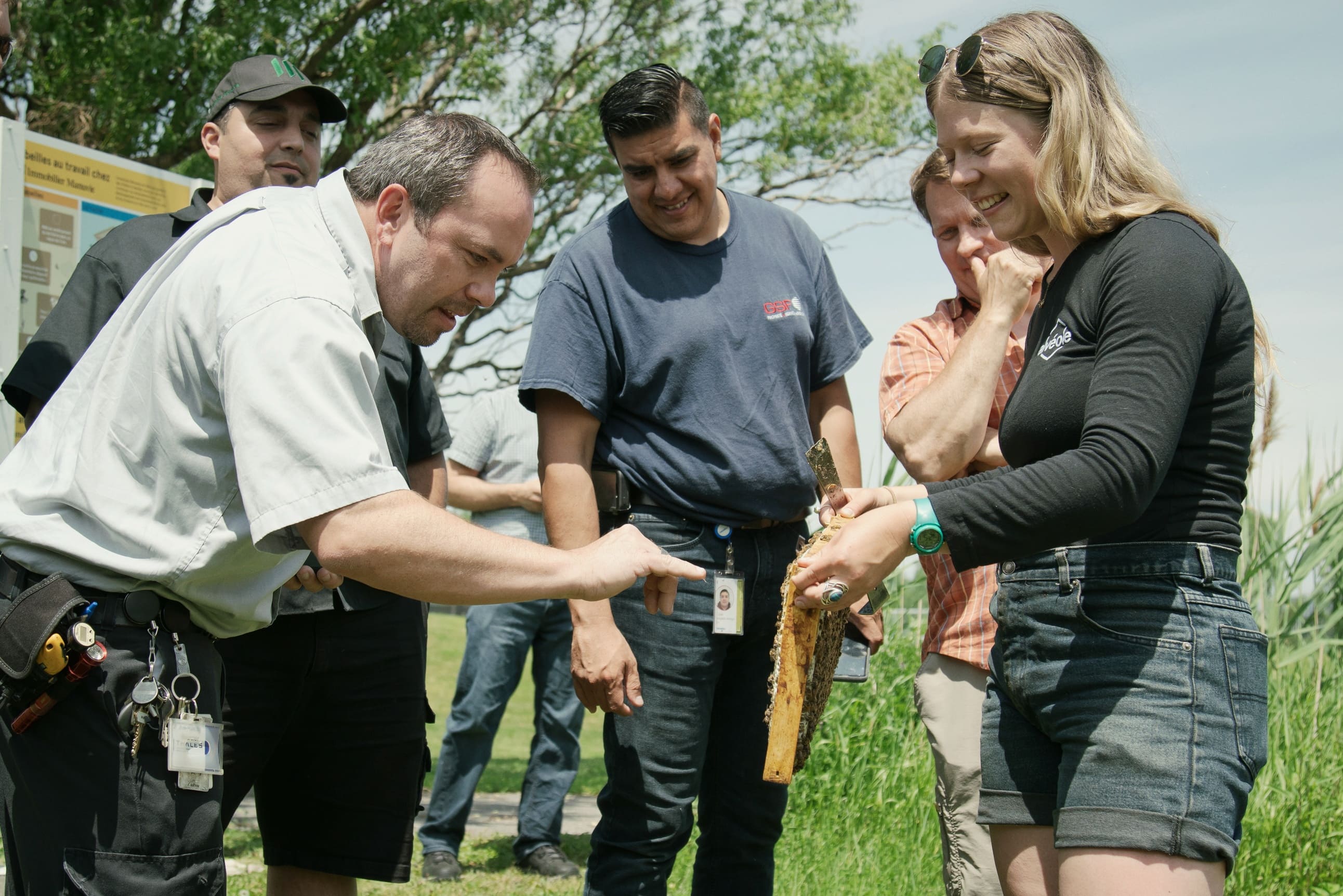 A group of people examining a honeycomb frame held by an Alveole beekeeper during an outdoor workshop