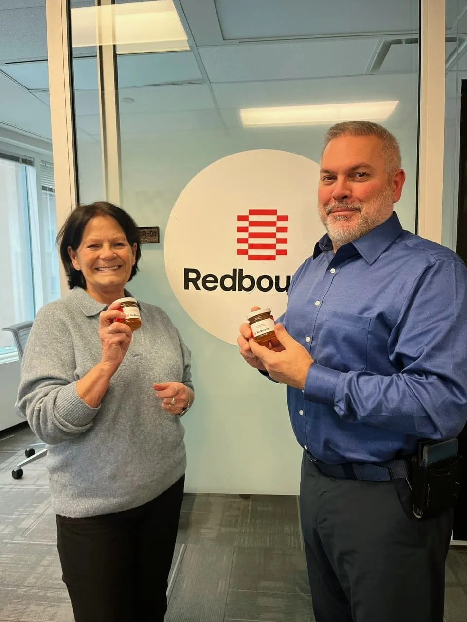 Two people holding branded Alveole honey jars in front of a Redbourne office logo