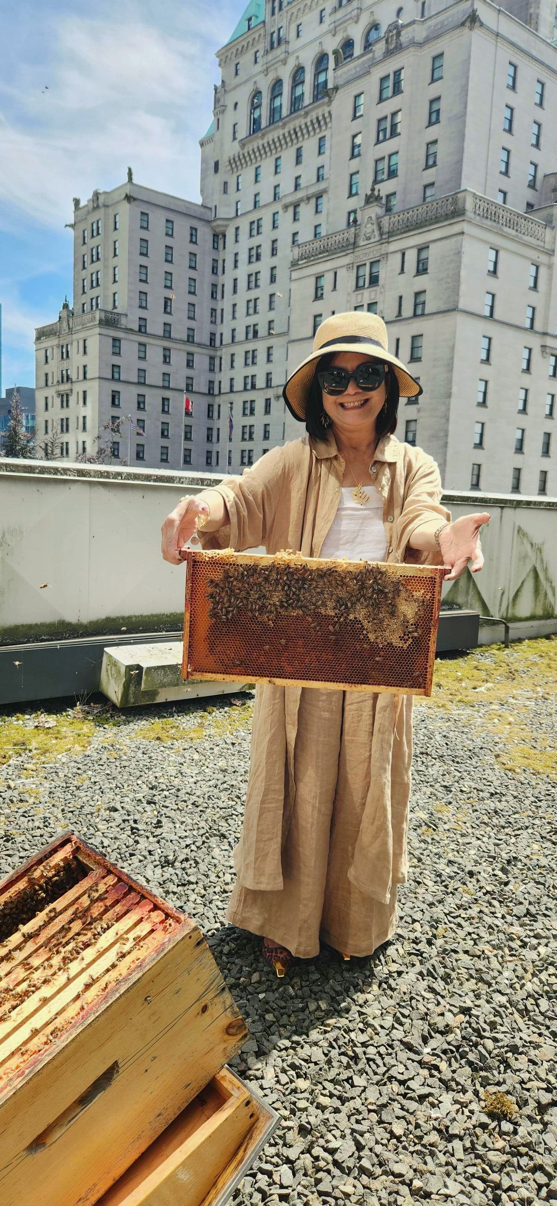 A woman smiling on a rooftop, holding a large honeycomb frame with bees, with a historic building behind her