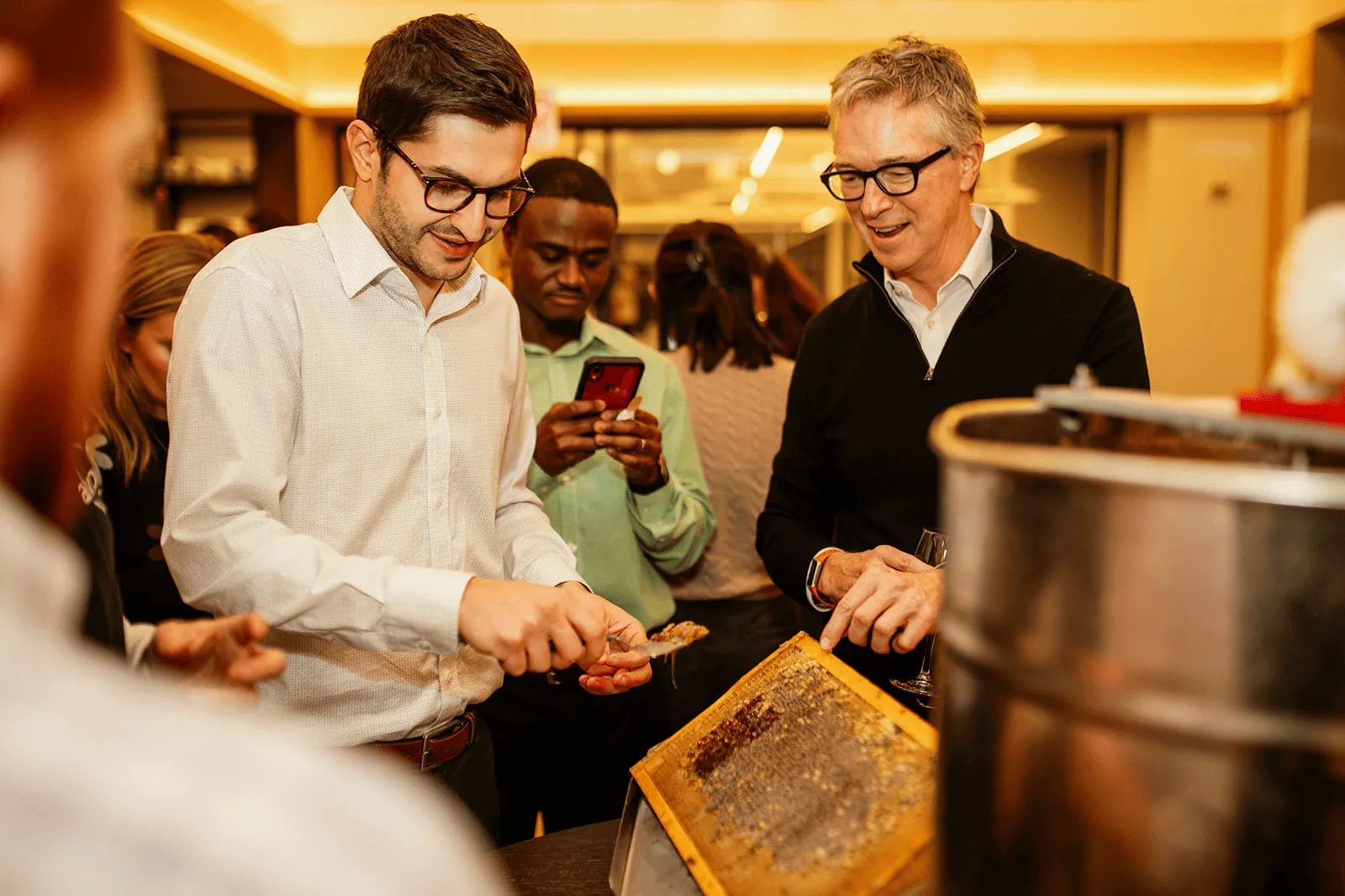 Two men at an indoor corporate event examining a honeycomb frame next to a honey extractor