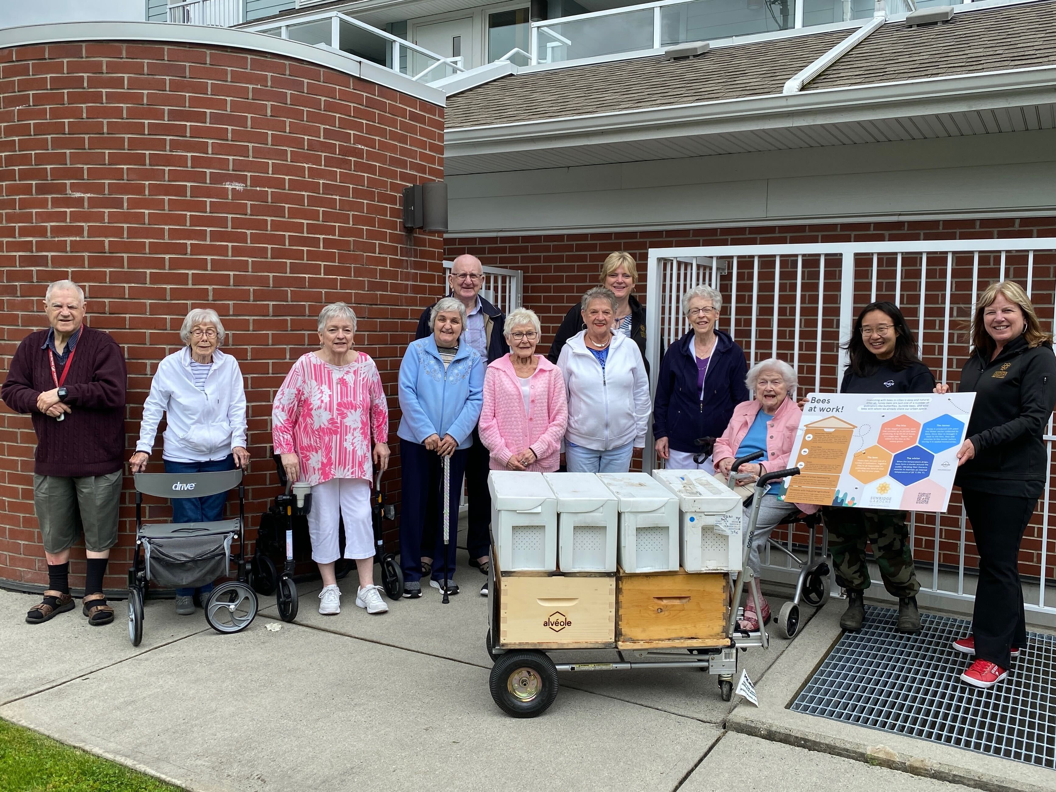 Senior residents and Alvéole staff gathered outdoors around a beehive on a cart, holding an educational poster about bees