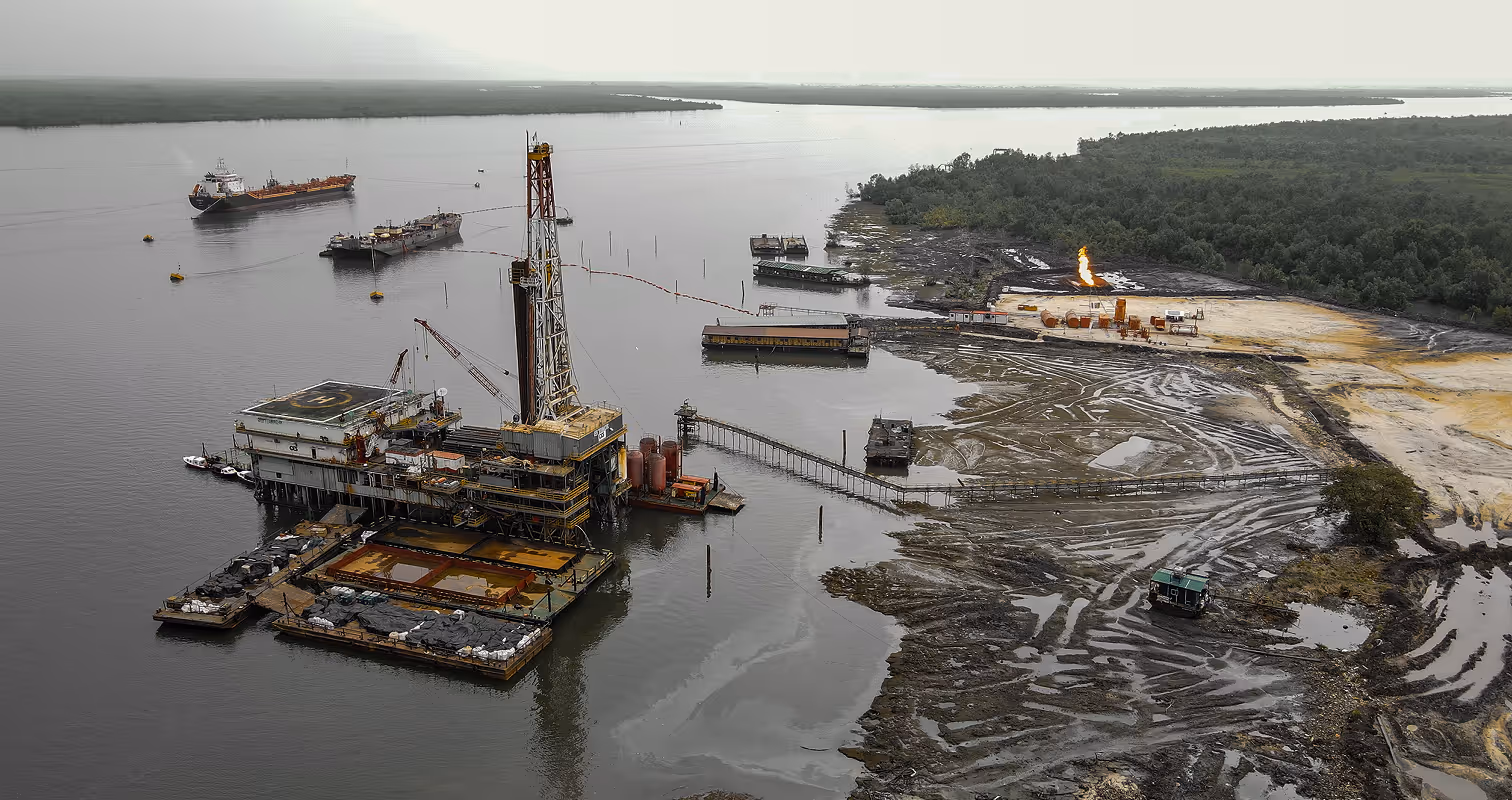 Offshore oil drilling platform connected to muddy shore with nearby ships and a flaming gas flare in the background.