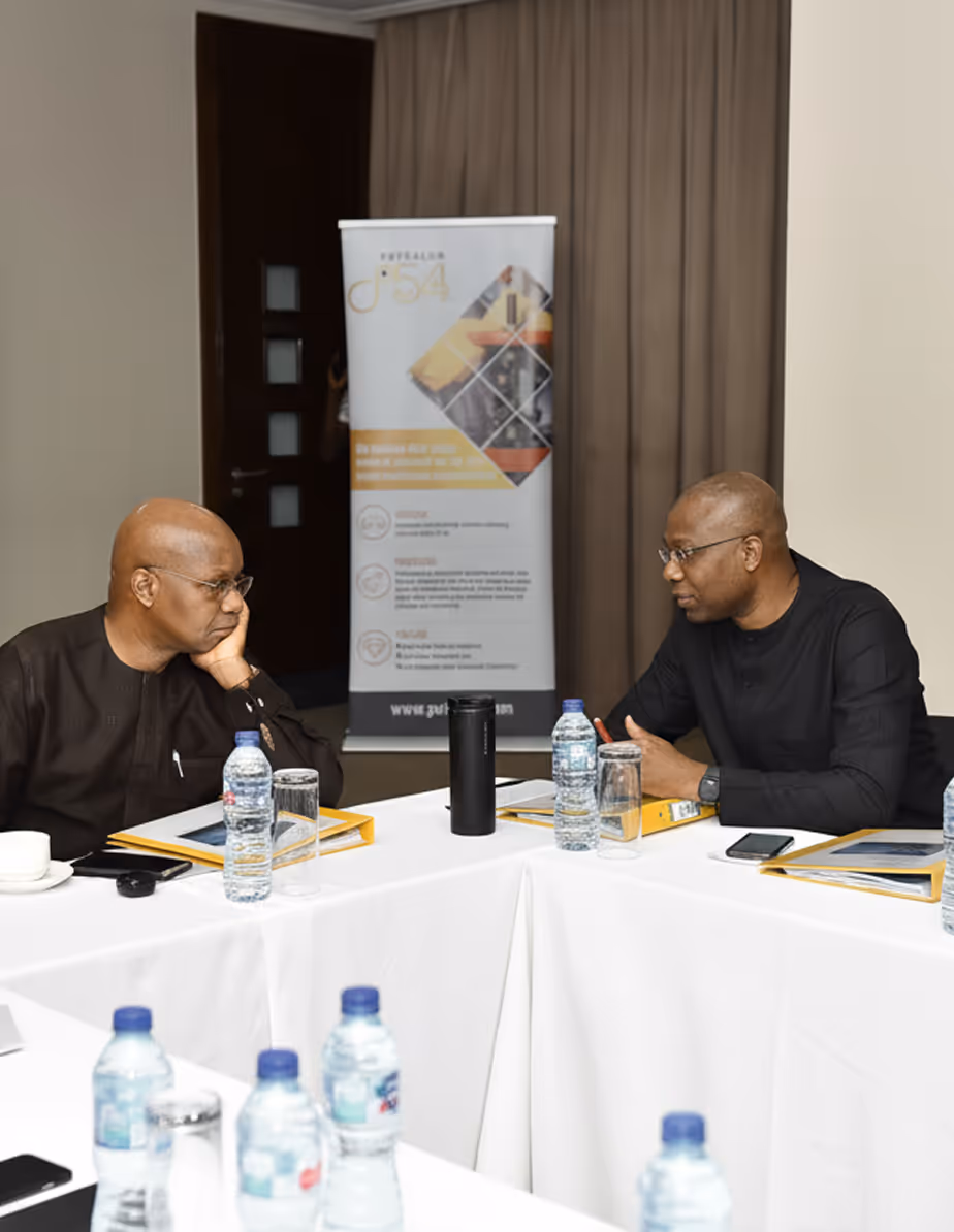 Two men in dark clothing sitting at a white conference table with water bottles, engaged in conversation.