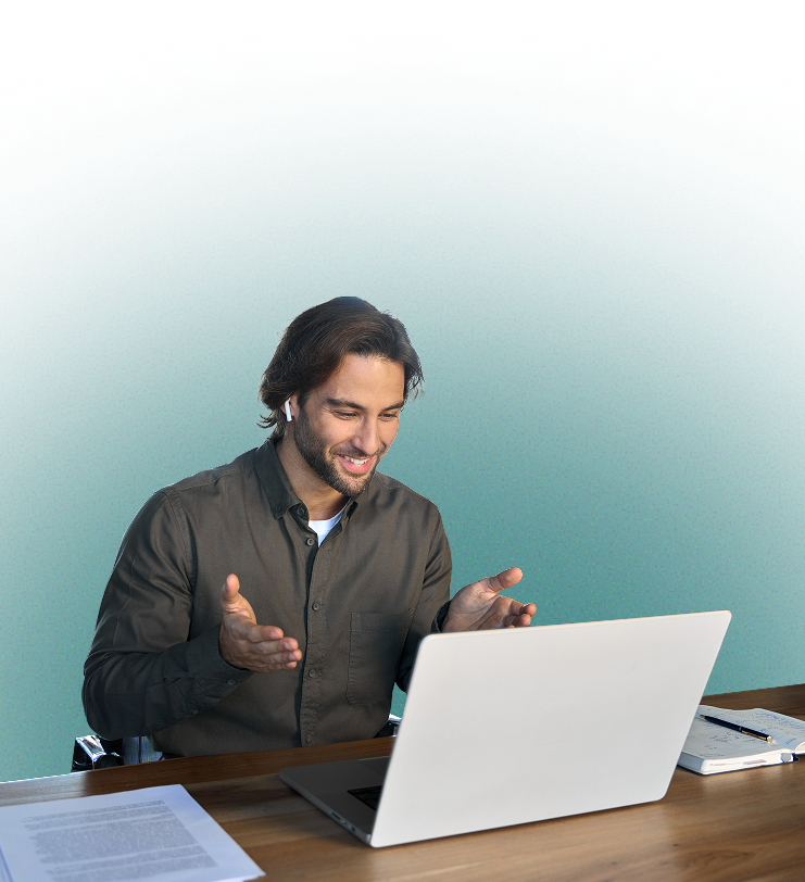 Smiling man wearing earbuds, gesturing while looking at a laptop on a wooden desk with papers and a notebook.