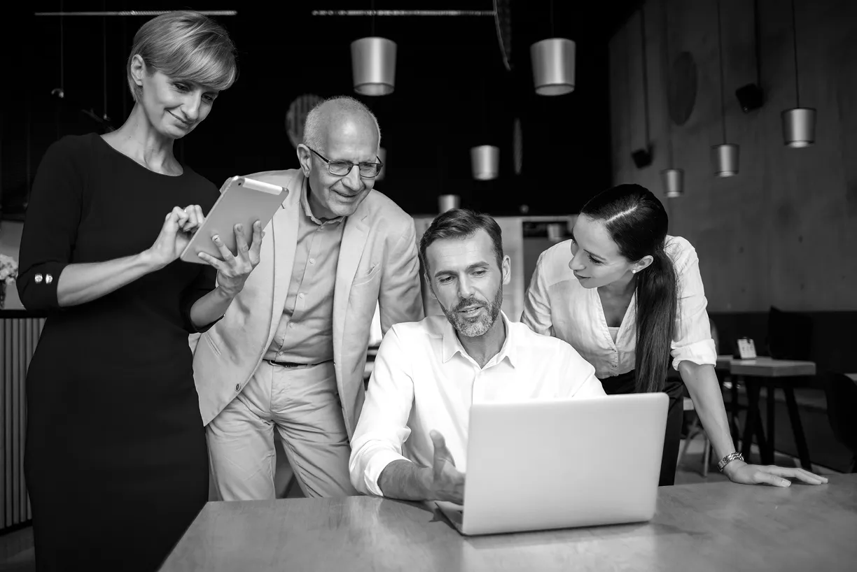 Four business colleagues collaborating around a laptop and tablet in a modern office setting.