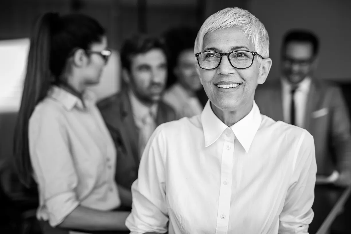 Smiling confident woman with short gray hair and glasses in the foreground with three business colleagues in the background in an office.