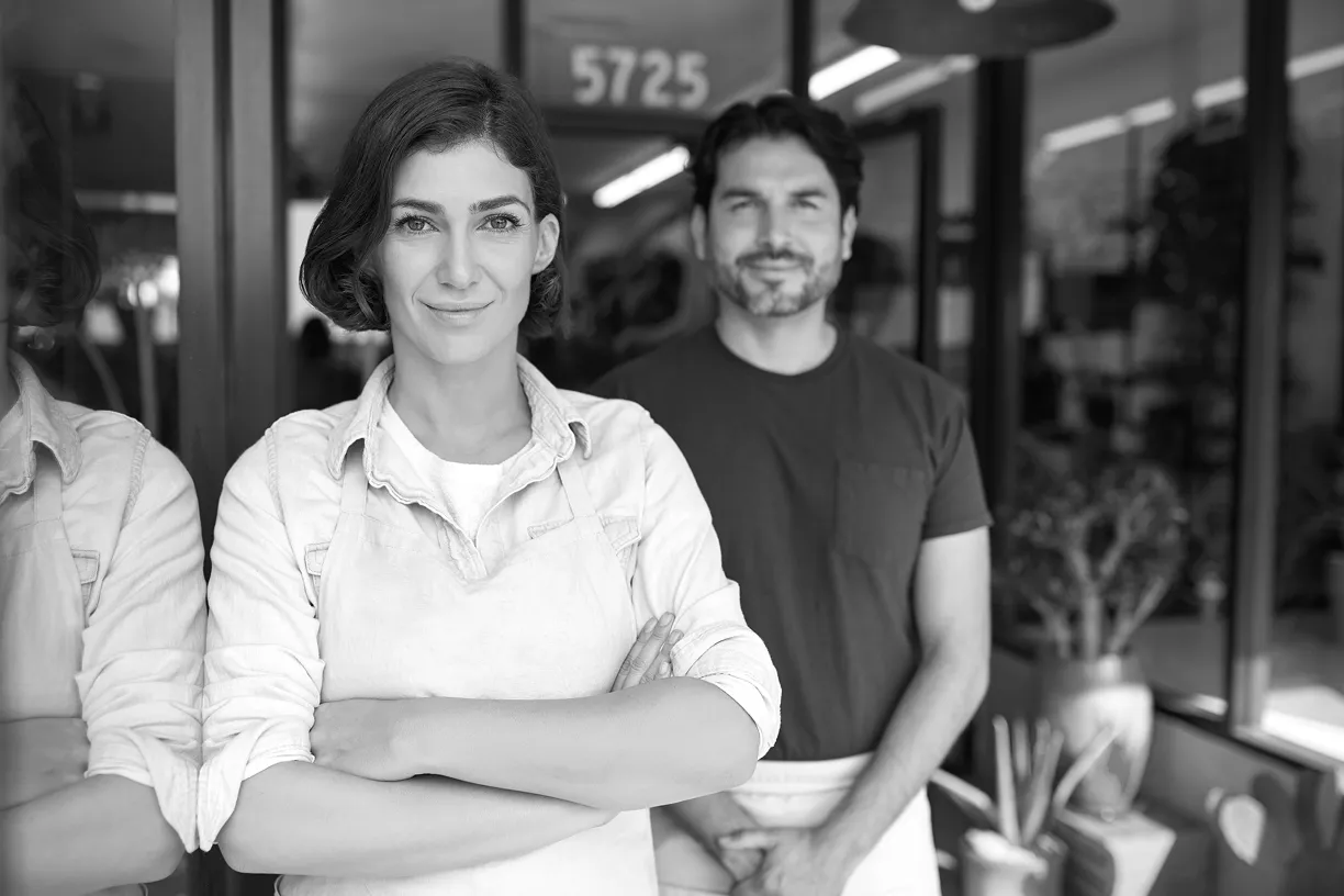 Confident woman in an apron stands with arms crossed in front of a man wearing an apron inside a shop.