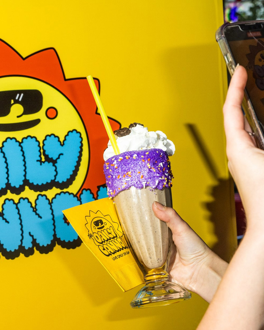 Hand holding a chocolate milkshake with whipped cream, purple sugar rim, and yellow straw in front of a bright yellow wall with colorful cartoon-style logo.