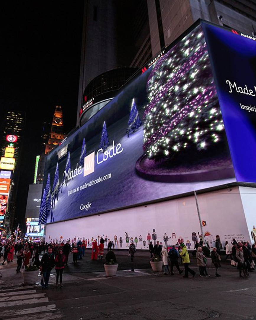 Large illuminated billboard at night in a city showing decorated Christmas trees with the text 'Made with Code' and a website link, above a pedestrian area with people walking.