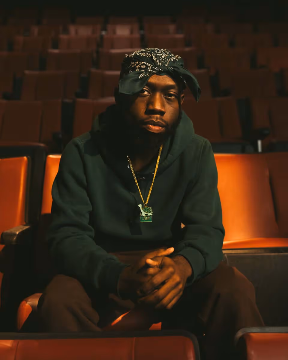 Man in black hoodie and bandana relaxing across orange theater seats in a cinematic portrait shot