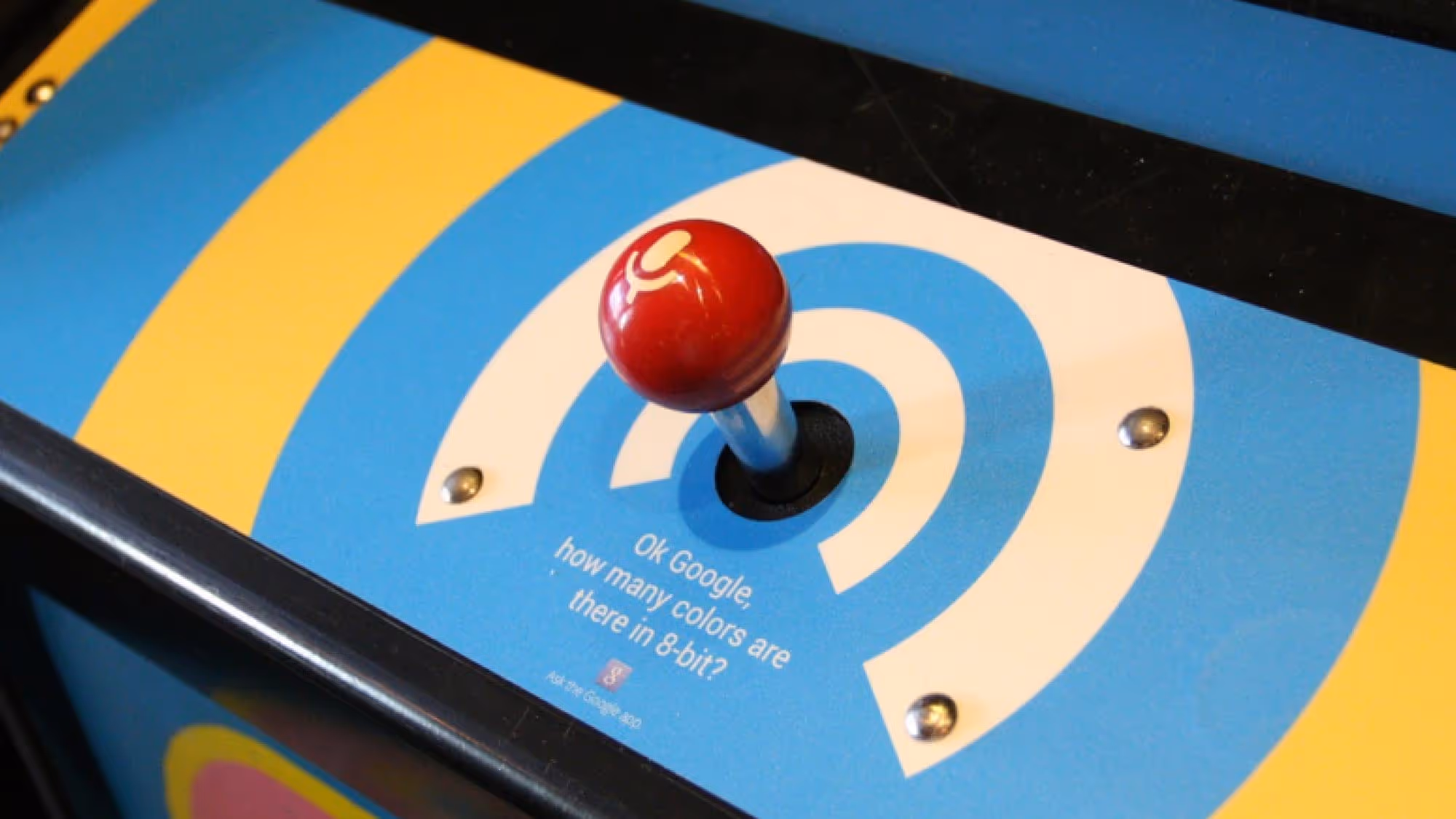 Vintage arcade joystick with red ball top on blue and yellow striped game panel asking 'OK Google, how many colors are there in 8-bit?