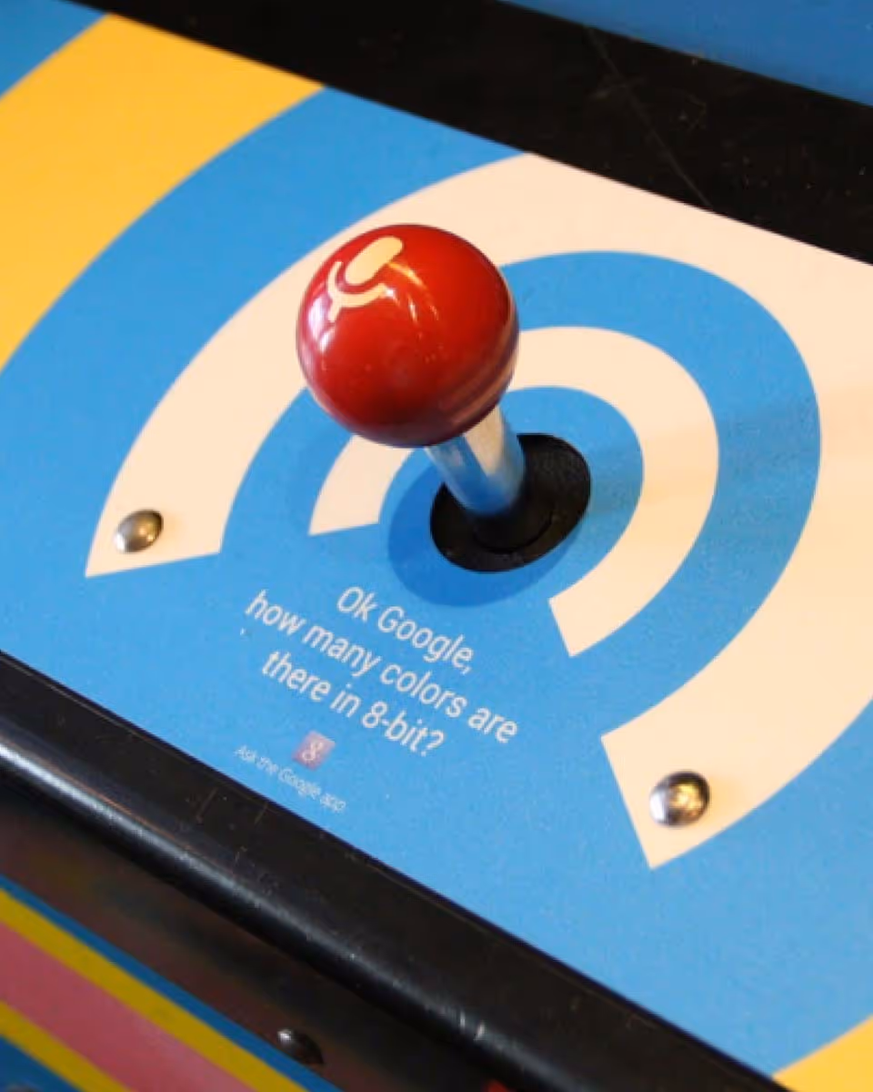 Vintage arcade joystick with red ball top on blue and yellow striped game panel asking 'OK Google, how many colors are there in 8-bit?