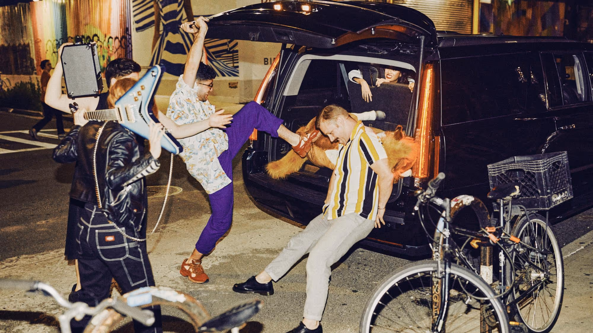 Behind-the-scenes photo shoot with people filling the trunk of a black SUV with music equipment and a stuffed horse on urban street at night
