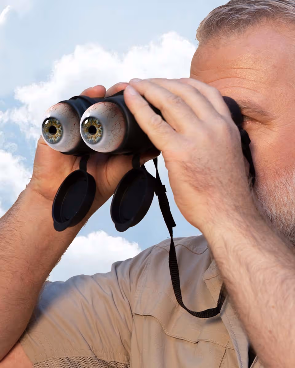 Man looking through binoculars with oversized googly eyes attached to the lenses against blue sky background