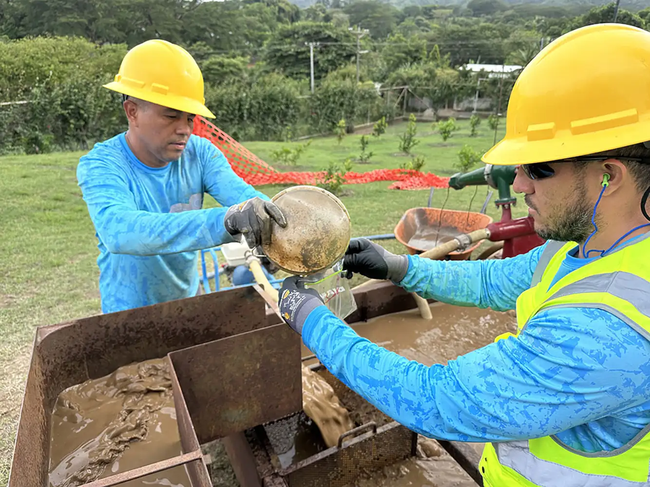 Two men on a water well drilling construction site in Nicaragua, testing water for HydroLOGICA. They are wearing blue shirts and yellow hard hats, with one in a bright yellow safety vest.