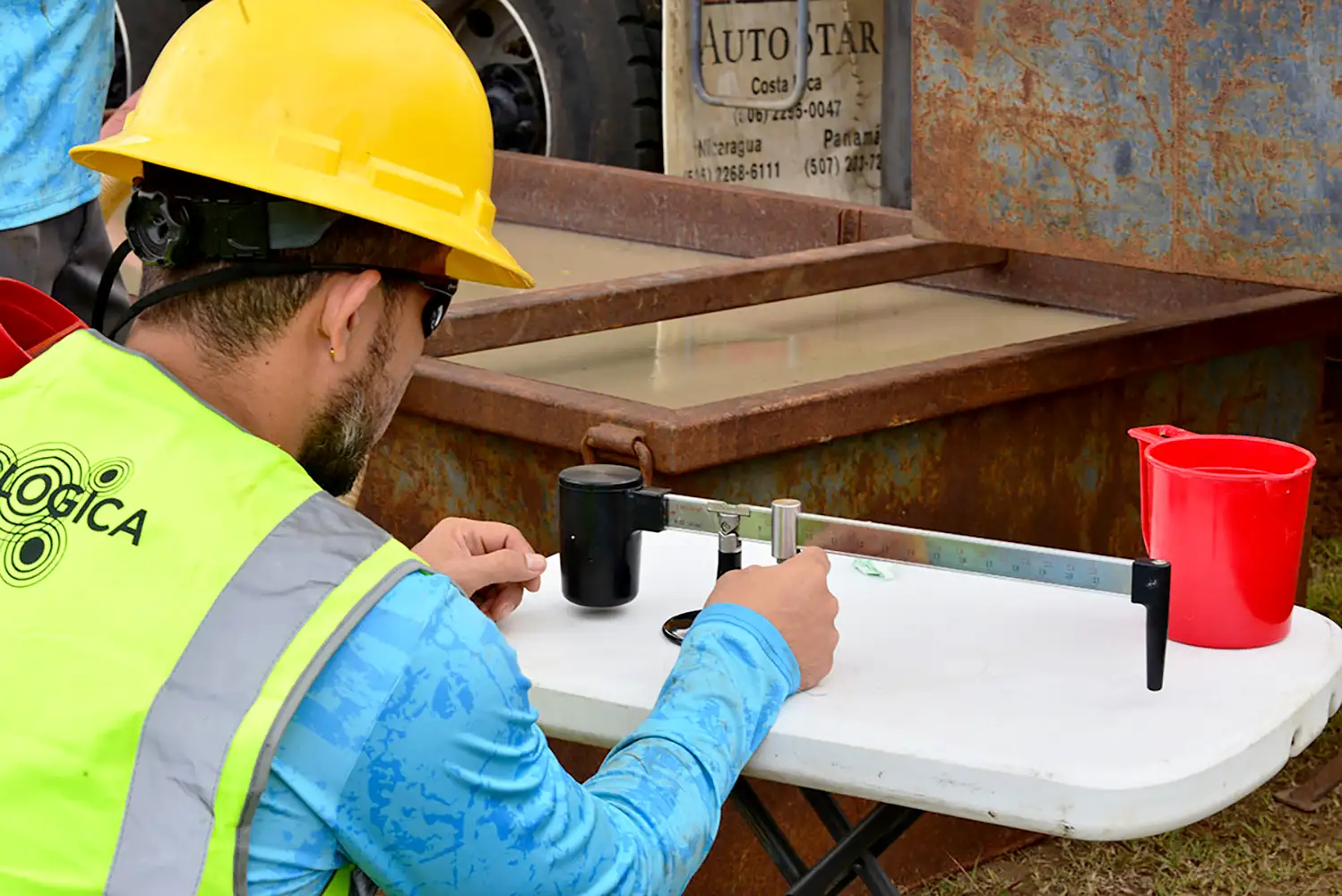 Collecting samples and measuring on site in Nicaragua, for HyrdoLOGICA. He is wearing a blue HydroLOGICA shirt, yellow safety vest and hard hat. He uses a weighted scale with focus.