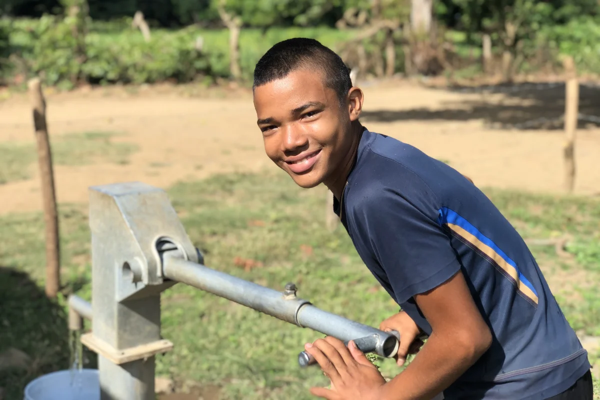 A child at a water pump, grinning.