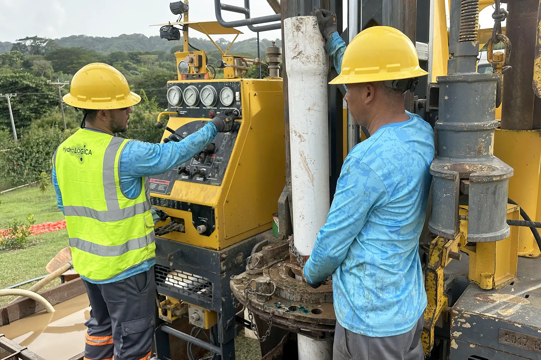 Two men on a construction site in Nicaragua, operating a water well drill for HydroLOGICA. They are wearing blue shirts and yellow hard hats, with one in a bright yellow safety vest.