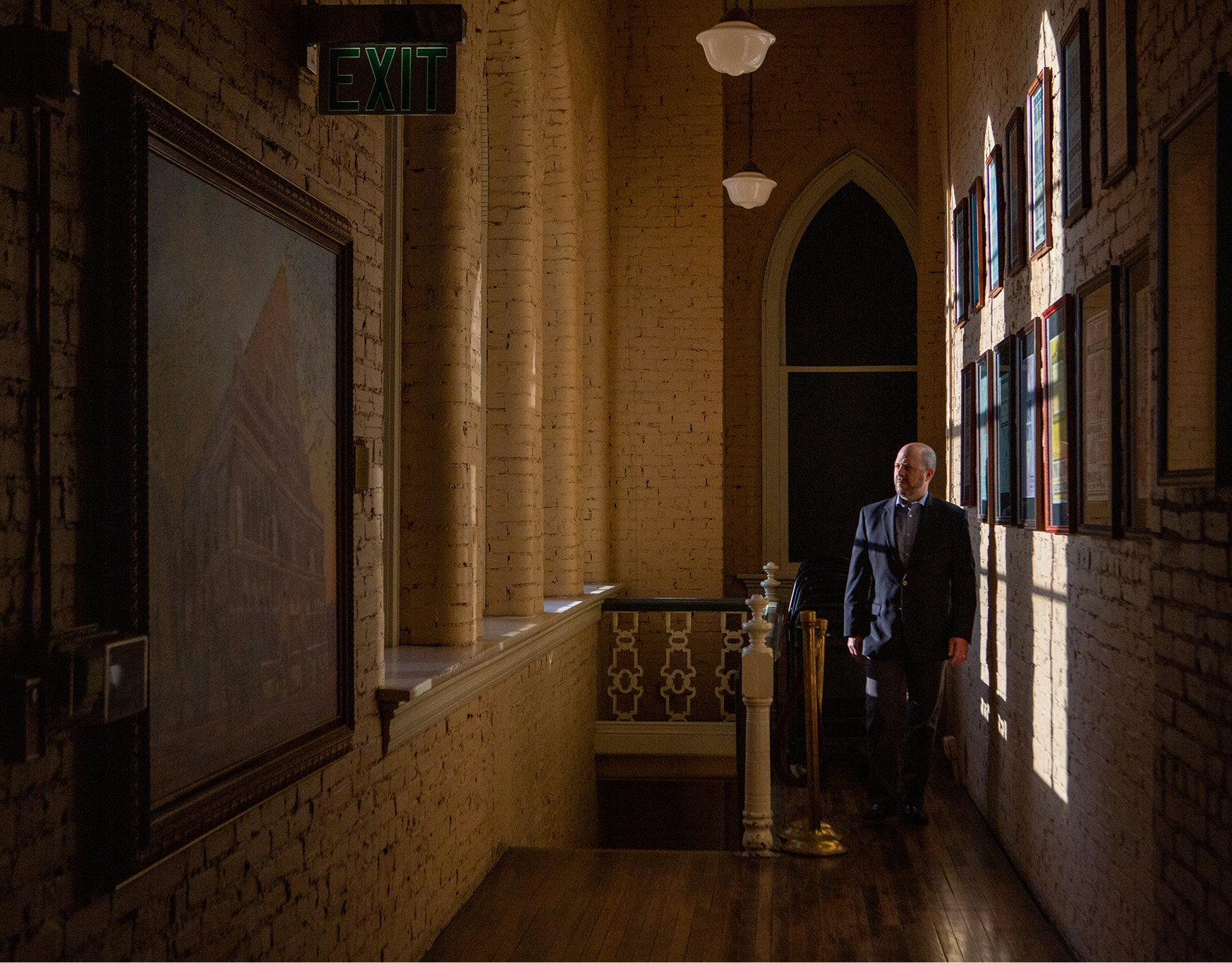 Man in a suit walking in a dimly lit hallway with brick walls and framed pictures illuminated by sunlight.