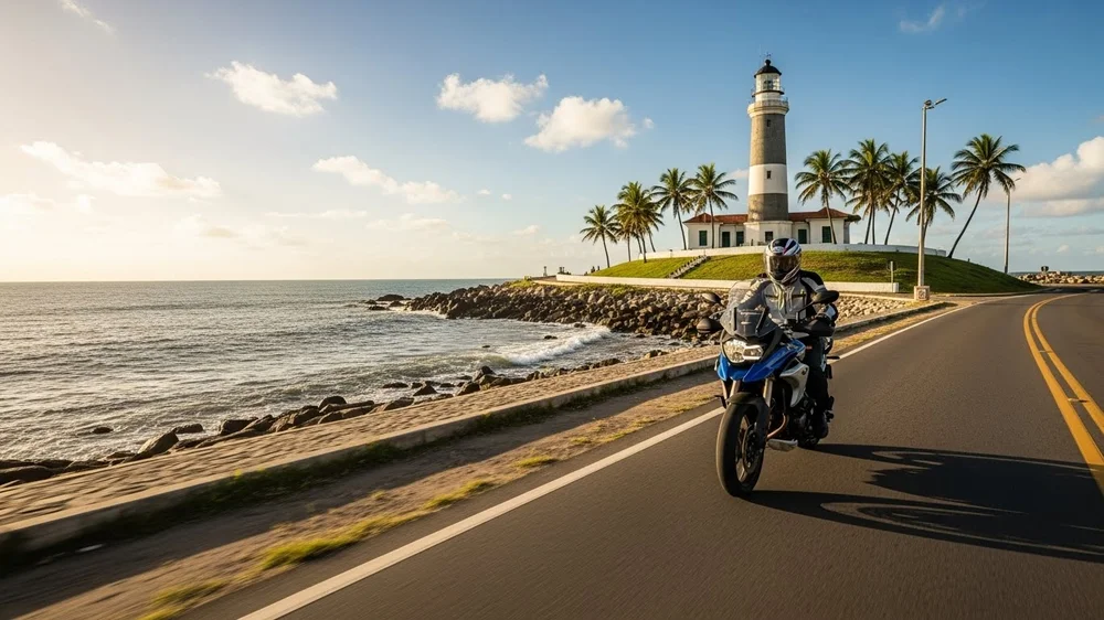 Motociclista em rodovia litorânea passando por farol histórico ao pôr do sol.