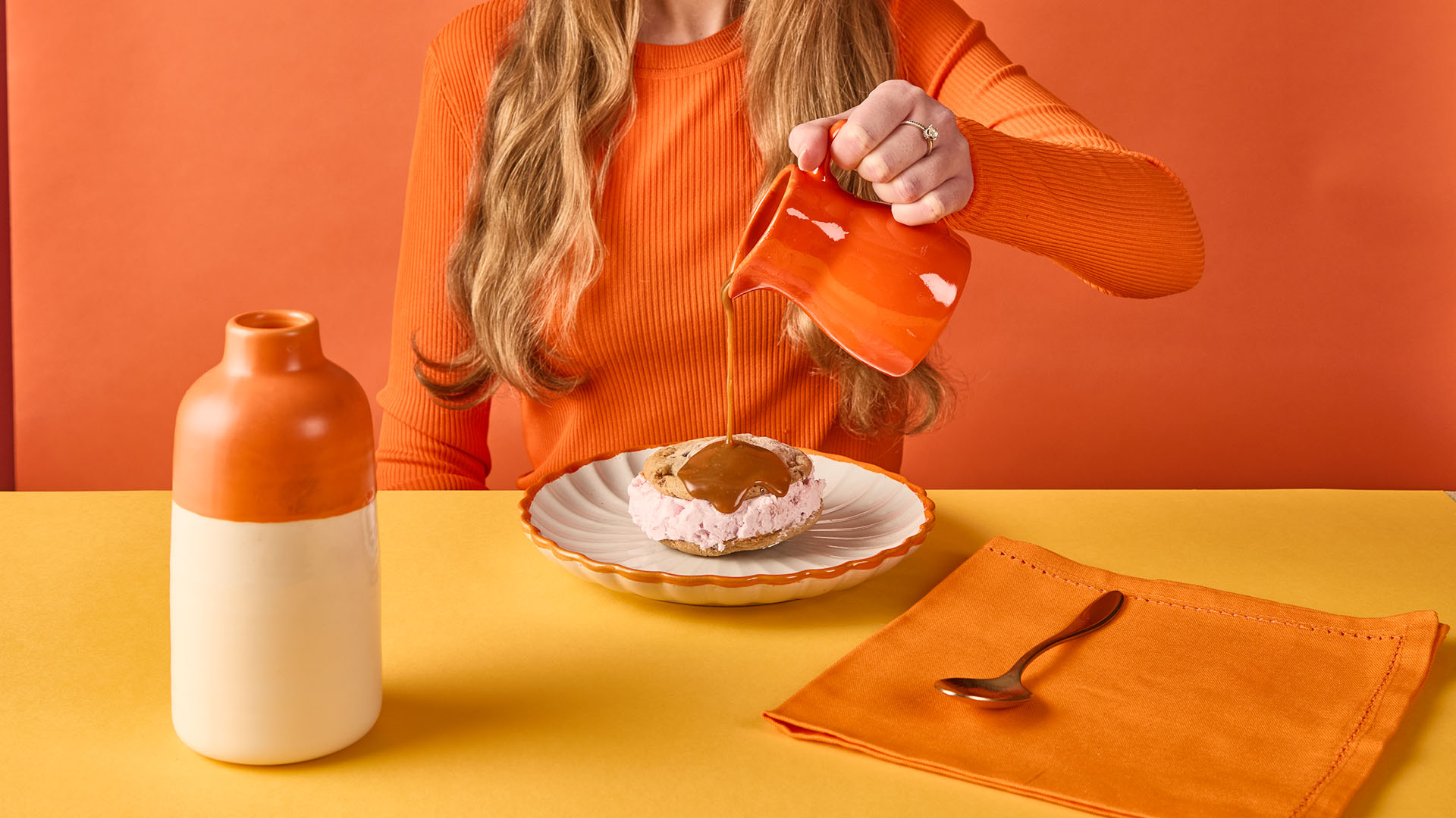 Person in an orange sweater pouring caramel sauce from an orange pitcher onto a cookie ice cream sandwich on a white plate with an orange rim.