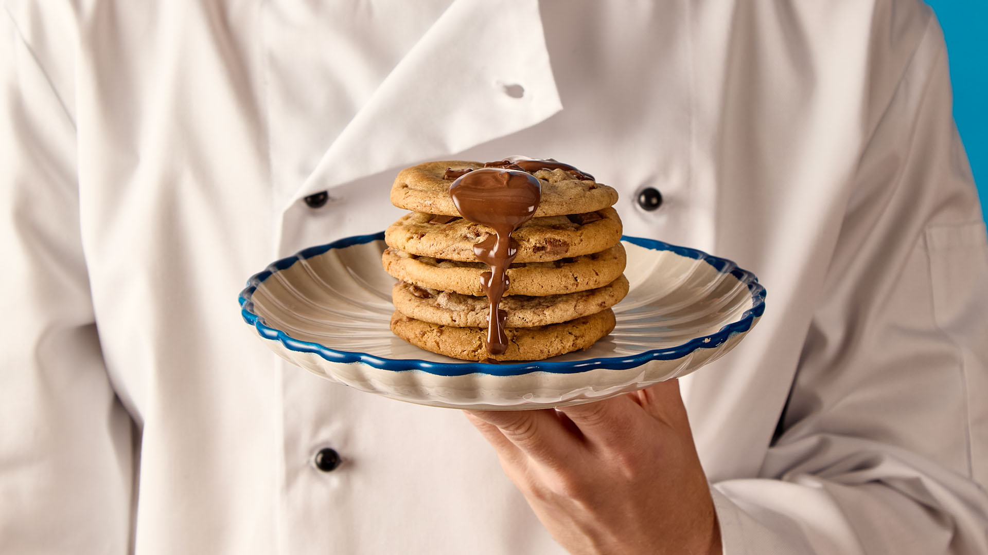 Stack of chocolate chip cookies with melted chocolate drizzle on a plate held by a person wearing a white chef coat.