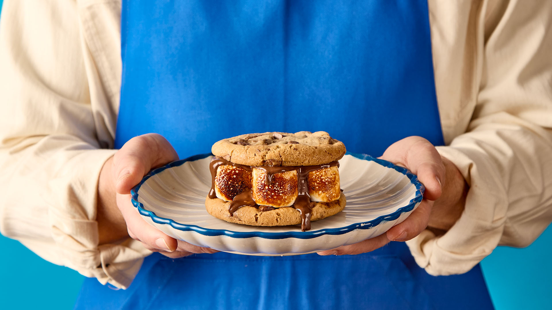 Hands holding a plate with a chocolate chip cookie sandwich filled with toasted marshmallows and melted chocolate.