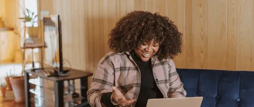 A Black girl with curly hair wearing a plaid shirt, laughing while looking at a tablet, sitting on a sofa.