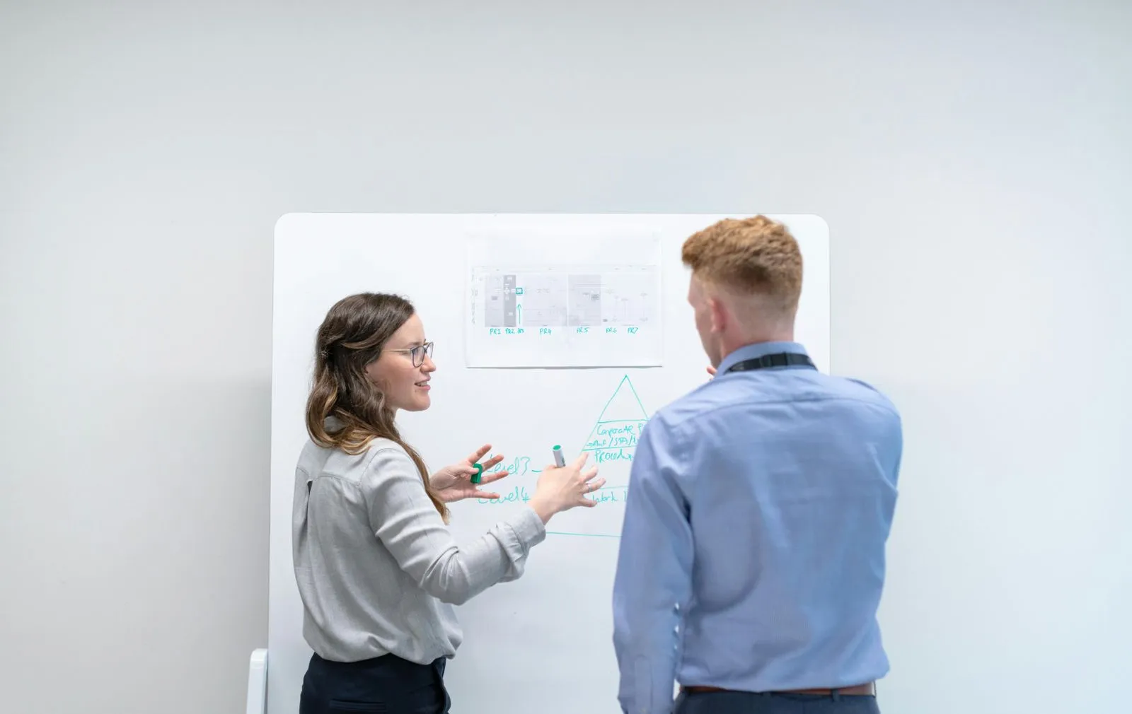 Two people in what appears to be a meeting with a gray wall and a chalkboard. There's a girl who seems to be explaining something to a guy at the whiteboard.