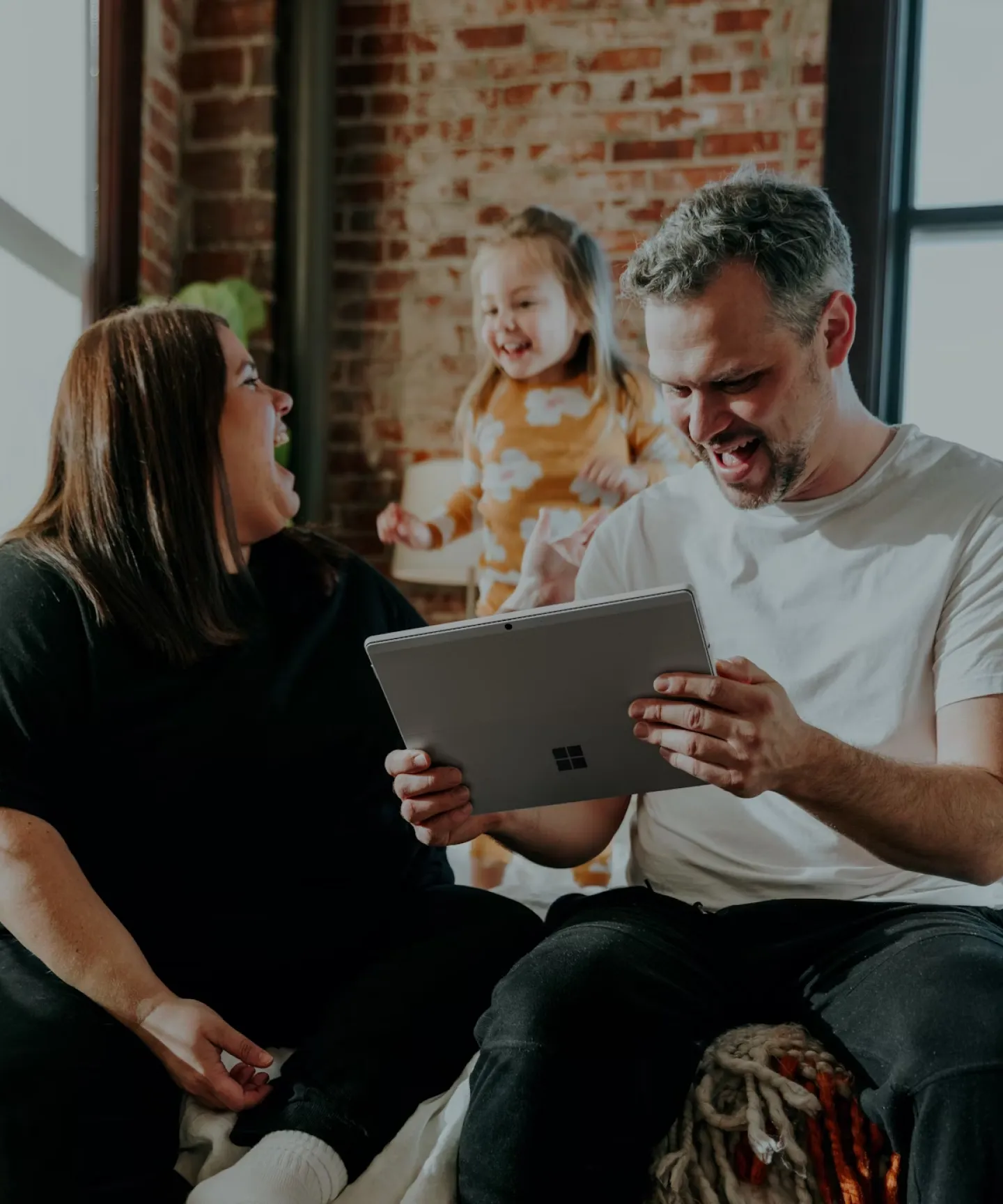 In the image, there are two parents and a girl. The father is holding a tablet and laughing at its content, while the mother and the girl are also laughing together.
