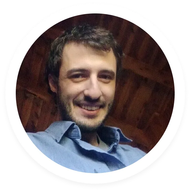 Smiling man with short dark hair and beard wearing a blue shirt inside a wooden-paneled room.