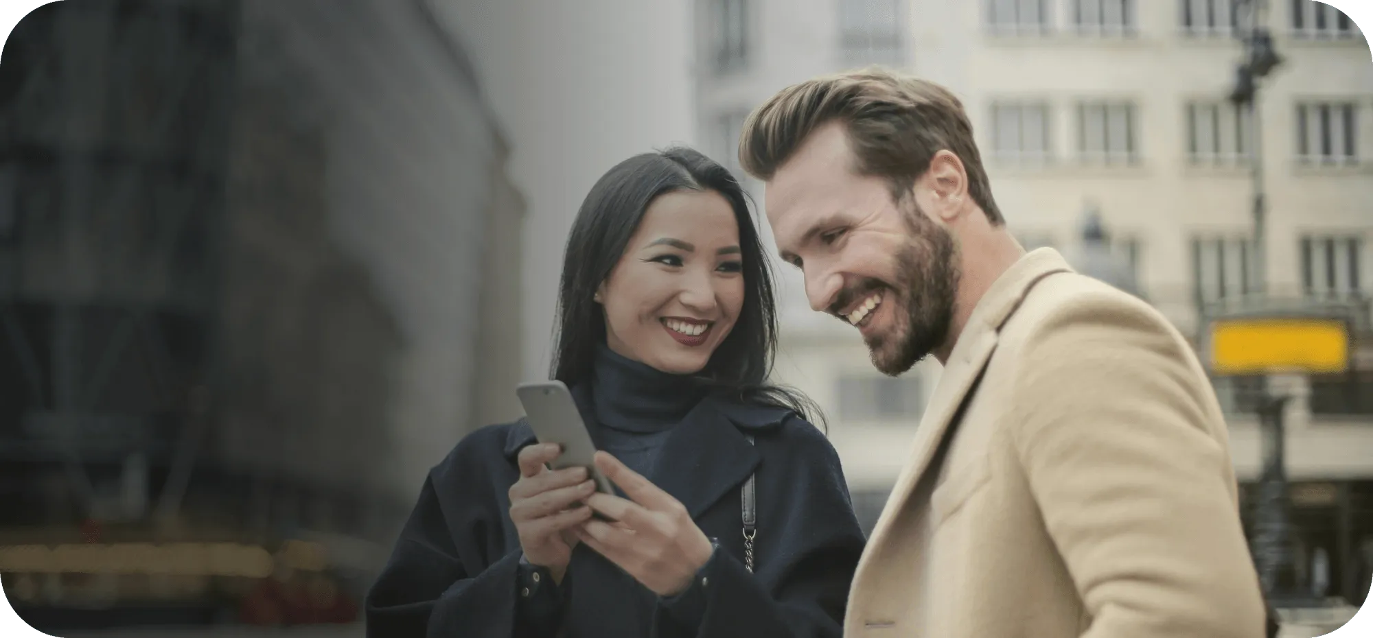 Smiling woman showing a smartphone to a laughing man outdoors in an urban setting.