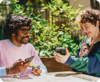 Two young people sitting outdoors at a table, smiling and looking at a smartphone together.