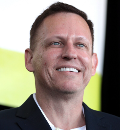 Smiling man with short brown hair wearing a dark blazer and white shirt against a blurred background.