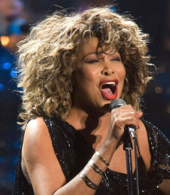 Woman with curly hair passionately singing into a microphone on stage, wearing a black sleeveless outfit and bracelets.