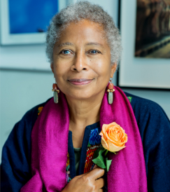 Older woman with gray curly hair wearing a magenta scarf and holding an orange rose, smiling gently.