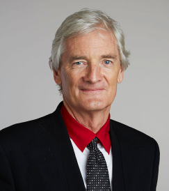 Portrait of an older man with gray hair wearing a black suit, red shirt, and black patterned tie against a gray background.