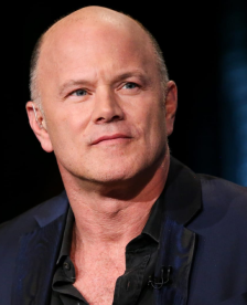 Bald man wearing a dark blazer and black shirt, looking slightly to the side against a dark background.