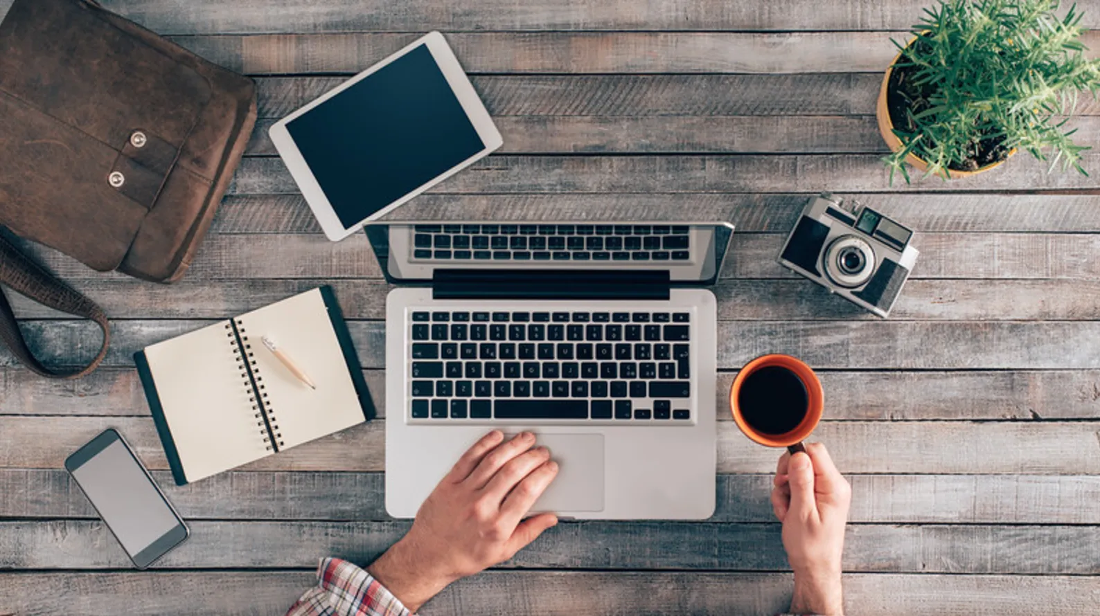 Person using a laptop at a wooden desk with a tablet, smartphone, notebook, camera, plant, and holding a cup of coffee.