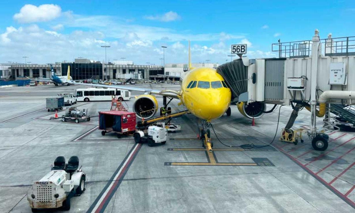 Yellow airplane parked at gate 56B with jet bridge connected and ground service vehicles nearby on a sunny day.