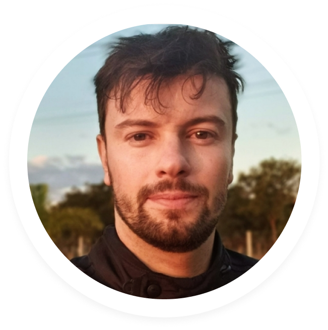 Close-up of a young man with short brown hair and beard, outdoors with trees in the background at sunset.