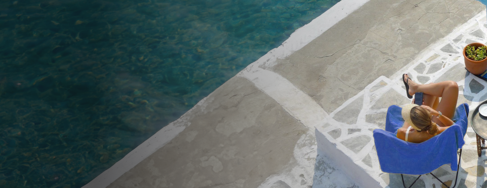 Woman sitting on a blue chair beside clear water, using a smartphone with legs crossed and wearing a straw hat.