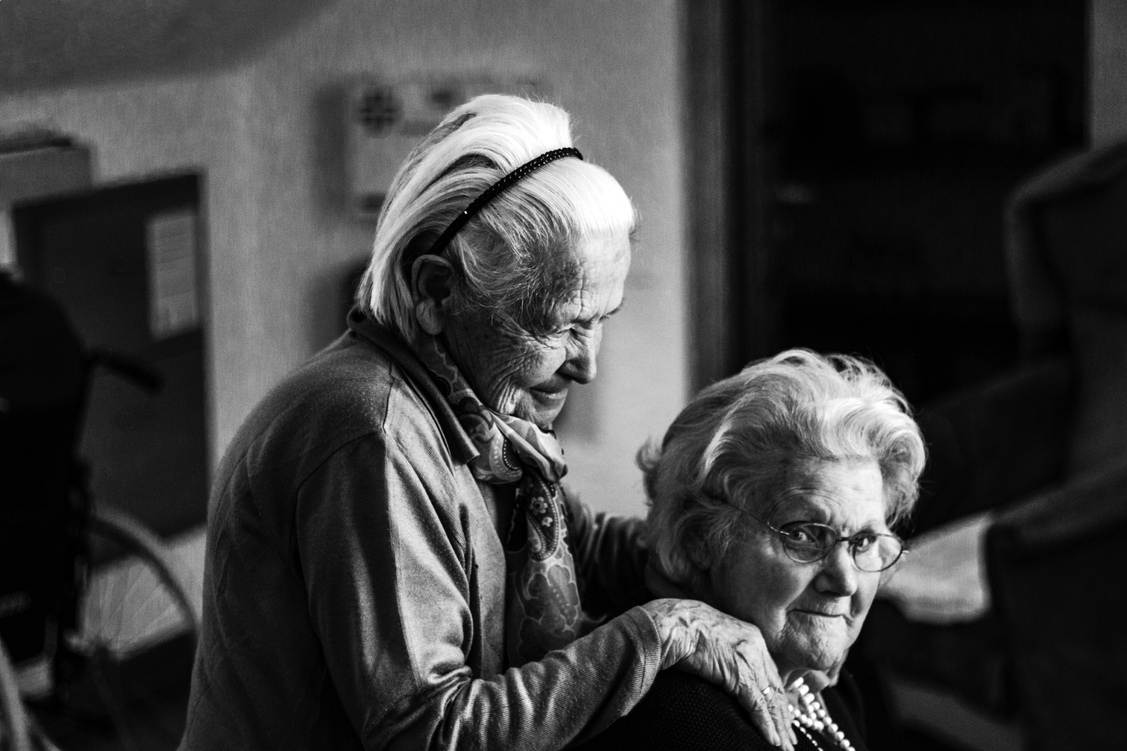 Black and white photo of two elderly women; one stands behind the other with gentle hands on her shoulders.